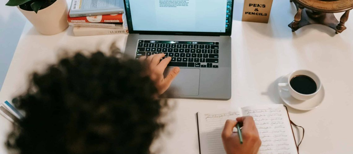 Person writing in notebook with laptop and coffee for writing prompts.